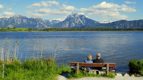 älteres Paar genießt die herrliche Aussicht von einer Bank über den Hopfensee auf die Berge im Allgäu 