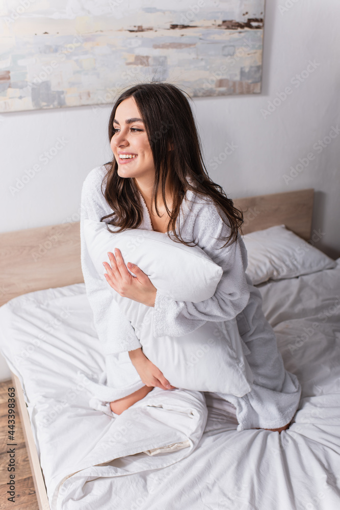 pleased woman in bathrobe holding pillow while sitting on bed pleased woman in bathrobe holding pillow while sitting on bed