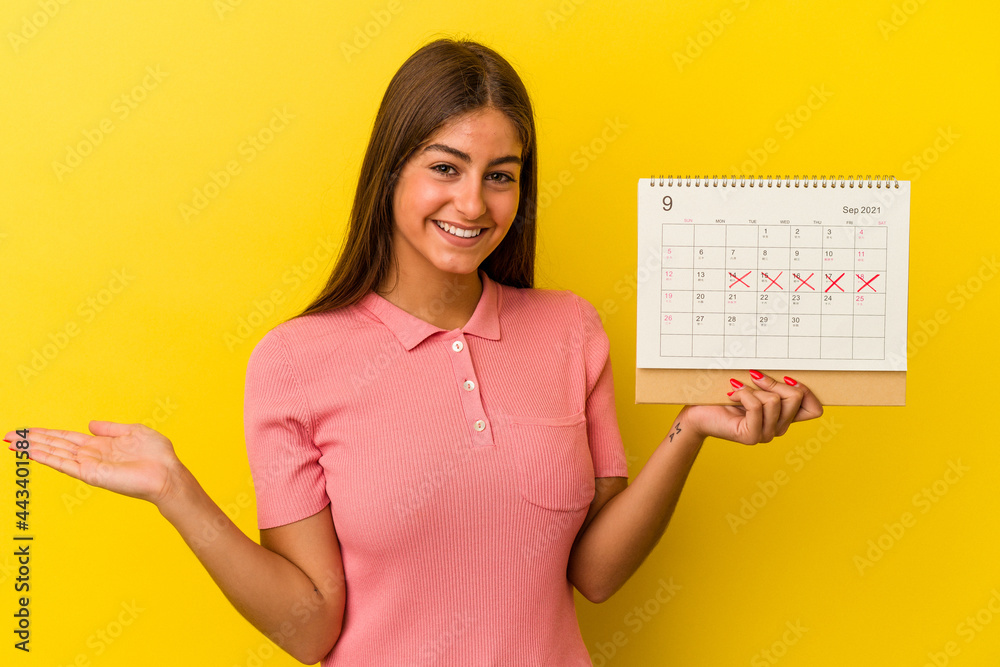 Young caucasian woman holding a calendar isolated on yellow background showing a copy space on a palm and holding another hand on waist. Young caucasian woman holding a calendar isolated on yellow background showing a copy space on a palm and holding another hand on waist.