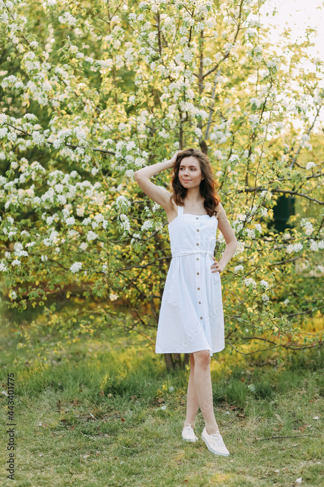 A close-up portrait of a happy pretty gentle smiling young woman with a hairstyle in a white cotton dress having fun walking alone enjoying the smell of blooming flowers in a summer green outdoor park A close-up portrait of a happy pretty gentle smiling young woman with a hairstyle in a white cotton dress having fun walking alone enjoying the smell of blooming flowers in a summer green outdoor park