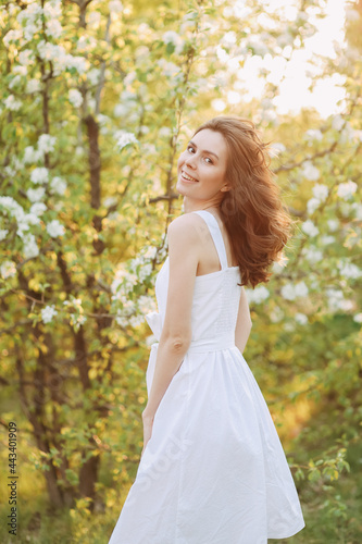 A close-up portrait of a happy pretty gentle smiling young woman with a hairstyle in a white cotton dress having fun walking alone enjoying the smell of blooming flowers in a summer green outdoor park
