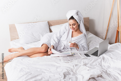 joyful woman in bathrobe and towel on head holding cup of coffee and reading newspaper near gadgets on bed
