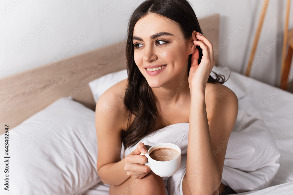 brunette woman smiling while adjusting hair and holding cup of coffee in bed brunette woman smiling while adjusting hair and holding cup of coffee in bed