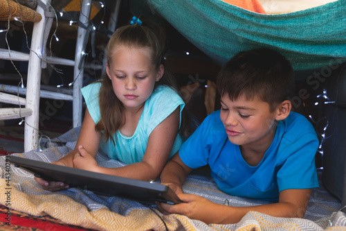 Caucasian brother and sister smiling, lying in blanket fort using tablet at home