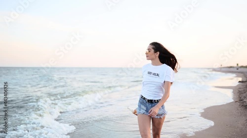 Pretty woman walking along seaside on sandy beach having fun with beer bottle. Attractive fitted girl raises hands, enjoys fredom and summer vacation near ocean shore, drinking beverage. Slow motion. photo