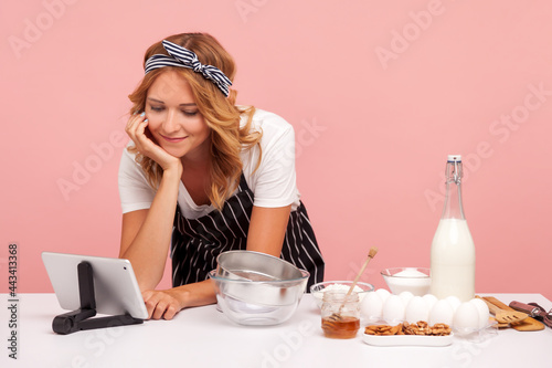Portrait of young adult female chef wearing pron using tablet for learning new recipe of pastry, looking at digital gadget and keeps hand under chin. Indoor studio shot isolated on pink background. photo
