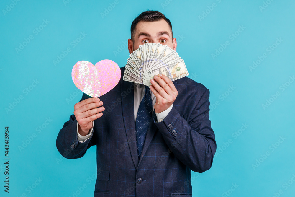 Portrait of lucky rich bearded man in official style suit hiding half of face with dollar banknotes and holding pink paper heart, enjoying wealth. Indoor studio shot isolated on blue background. Portrait of lucky rich bearded man in official style suit hiding half of face with dollar banknotes and holding pink paper heart, enjoying wealth. Indoor studio shot isolated on blue background.
