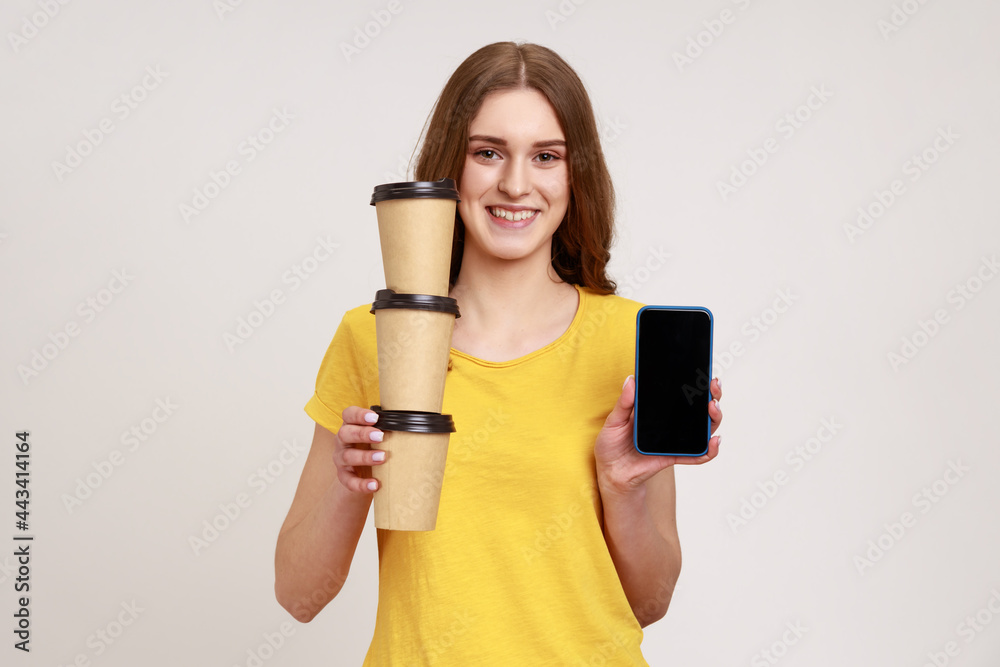 Delivery mobile app, internet advertise. Happy friendly teenager girl in yellow T-shirt holding stack of coffee cups and cellphone with mock up display. Indoor studio shot isolated on gray background. Delivery mobile app, internet advertise. Happy friendly teenager girl in yellow T-shirt holding stack of coffee cups and cellphone with mock up display. Indoor studio shot isolated on gray background.
