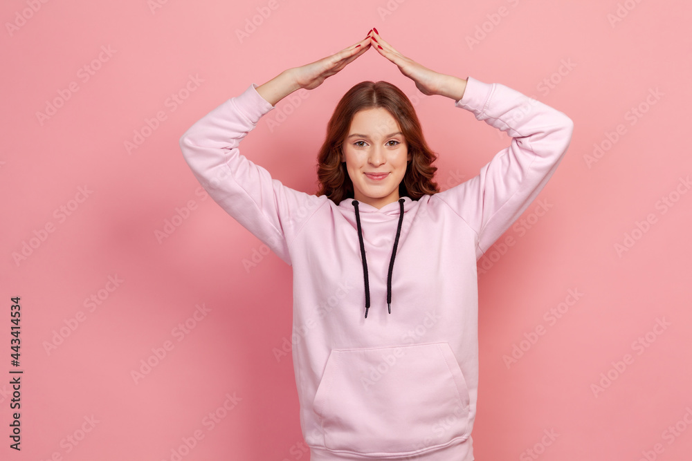 Portrait of friendly curly haired teenager girl in hoodie showing house roof symbol over head and smiling, feeling secured. Indoor studio shot isolated on pink background