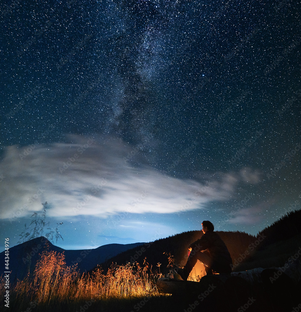 Fototapeta premium Side view of silhouette of man relaxing near forest and watching night sky with stars and Milky way. Male tourist is sitting near bonfire in mountains in evening. Concept of travelling.
