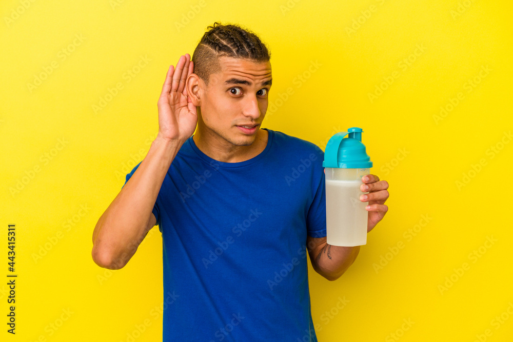 Young venezuelan man drinking a protein shake isolated on yellow background trying to listening a gossip. Young venezuelan man drinking a protein shake isolated on yellow background trying to listening a gossip.