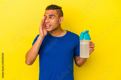 Young venezuelan man drinking a protein shake isolated on yellow background shouting and holding palm near opened mouth. © Asier