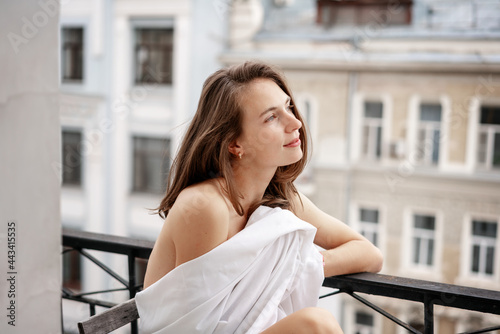 Beautiful young sensual woman with long dark hair wrapped in a white sheet sitting on the balcony and looking at the city