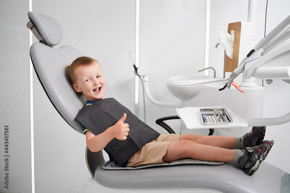 Horizontal side view snapshot of a little smiling boy wearing black bib, sitting in dentist chair, thumb up. Well equipped dentist office. Children's dentistry and medicine concept Horizontal side view snapshot of a little smiling boy wearing black bib, sitting in dentist chair, thumb up. Well equipped dentist office. Children's dentistry and medicine concept