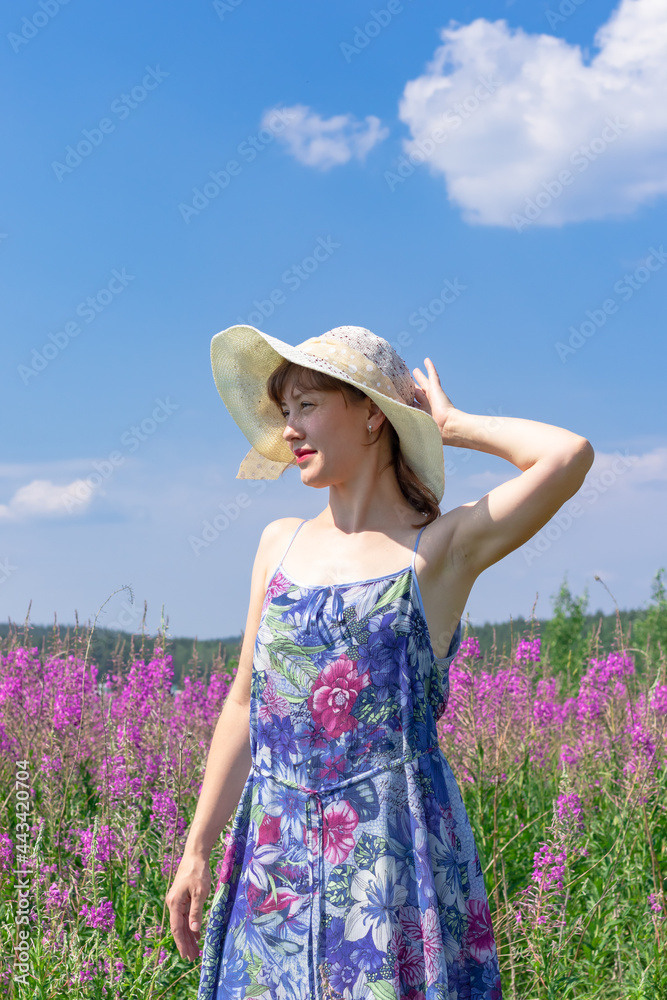 Young woman in a field of flowers in a straw hat against a background of green forest and blue sky on a hot summer day. Selective focus