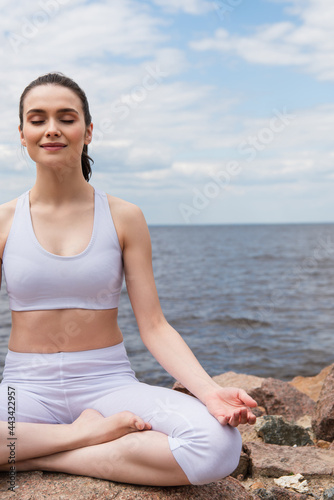cheerful woman in sportswear sitting in lotus pose while meditating near sea