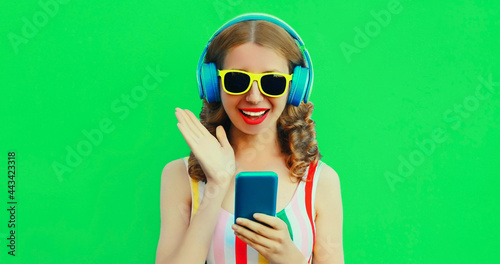 Summer colorful portrait of happy smiling young woman listening to music in headphones with phone on green background