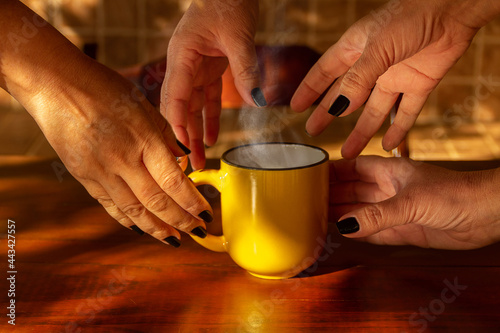 Quatro mãos, femininas, com unhas pintadas de preto, pegando uma caneca amarela com uma bebida quente.