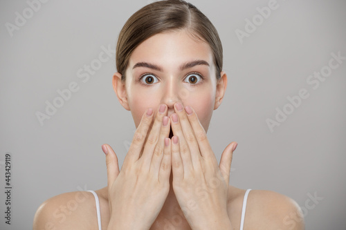 Beauty portrait. A beautiful girl with clean skin is surprised and covers her mouth with her hands. Gray background.