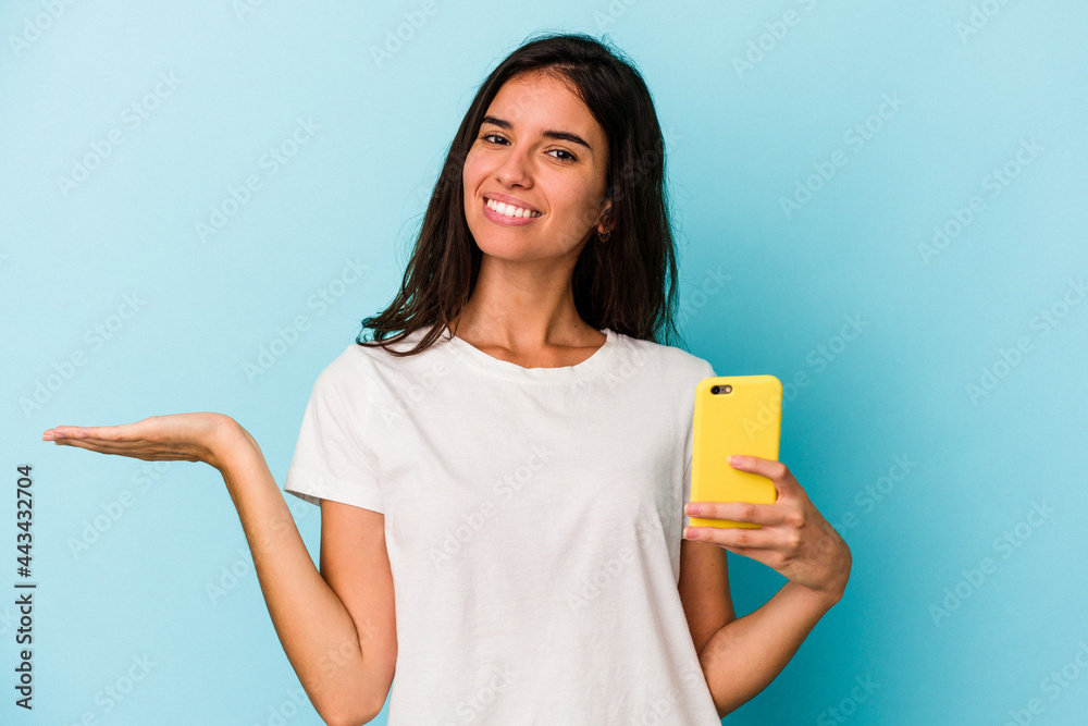 Young caucasian woman holding a mobile phone isolated on blue background showing a copy space on a palm and holding another hand on waist. Young caucasian woman holding a mobile phone isolated on blue background showing a copy space on a palm and holding another hand on waist.