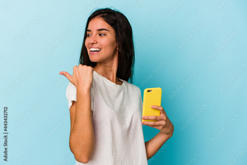 Young caucasian woman holding a mobile phone isolated on blue background points with thumb finger away, laughing and carefree. Young caucasian woman holding a mobile phone isolated on blue background points with thumb finger away, laughing and carefree.