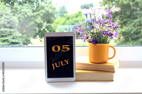 Calendar for July 5 : the name of the month of July in English and the numbers 0 and 5 on the tablet, a stack of books, wildflowers in a yellow cup on the background of the window