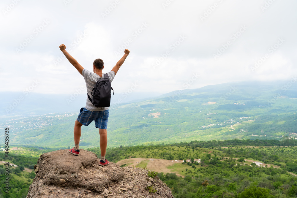 Young man standing on top of hill with hands up wearing gray backpack. Young guy raising hands up in nature.  Concept of travel lifestyle and vacation in mountains