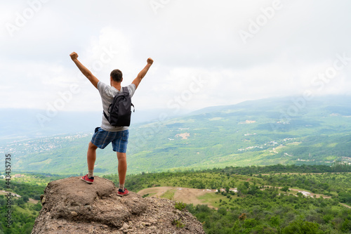Young man standing on top of hill with hands up wearing gray backpack. Young guy raising hands up in nature. Concept of travel lifestyle and vacation in mountains