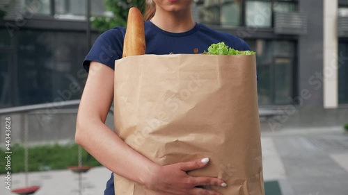 Close up food delivery woman courier holds paper bag with groceries vegetables bread in hand. Food delivery female worker employee in blue cap and t-shirt looking camera. Online internet shopping