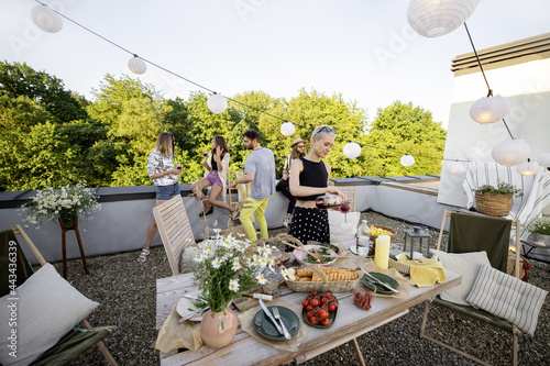 Young group of stylish people having a festive dinner on the beautifully decorated roof terrace. Friends hanging out together at picnic. Wide view