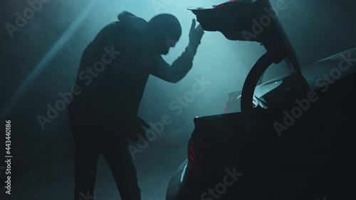 Handheld slowmo shot of young male criminal opening trunk of getaway car parked in dark garage He is looking into bag full of money and high-fiving his accomplice after successful robbery