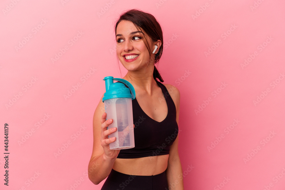 Young caucasian woman drinking a protein shake isolated on pink background looks aside smiling, cheerful and pleasant.