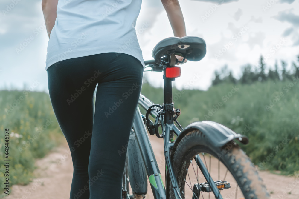 Obraz premium Toung slender blonde woman in a white shirt, black leggings rolls a bicycle on a sandy road in the middle of a field on a cloudy summer day, photo from the back