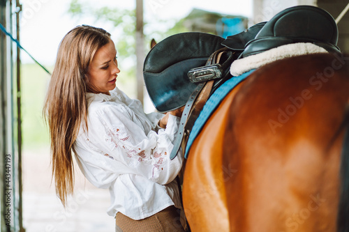 Young woman rider wearing a white shirt and hat with her brown horse in a stall, portrait. © Olga