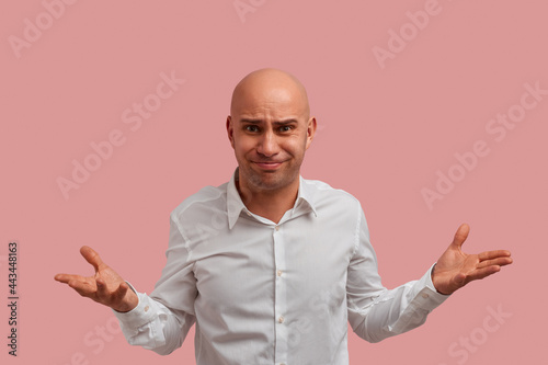 Why you do this. Horizontal shot of bald guy with bristle. With puzzled expression, spreads hands with confused look, frowns face, feels confused. Dressed in white shirt, isolated on pink background.