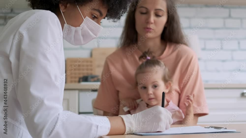 African american woman doctor in mask writing a prescription while examining a sick little girl. Family Doctor, Patient Support, Help at Home, Caring for the Sick. African american woman doctor in mask writing a prescription while examining a sick little girl. Family Doctor, Patient Support, Help at Home, Caring for the Sick.