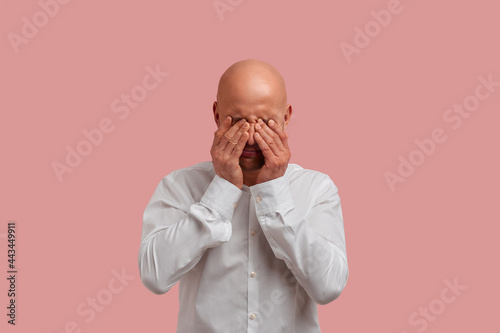Portrait of upset bald man with bristle in white shirt, rubs tears, feels badly with his problems. Posing in studio over pink background.