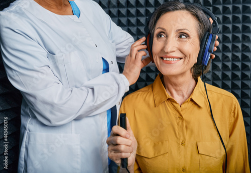 Portrait senior woman with white toothy smile while hearing check-up with ENT-doctor at soundproof audiometric booth using audiometry headphones and audiometer photo