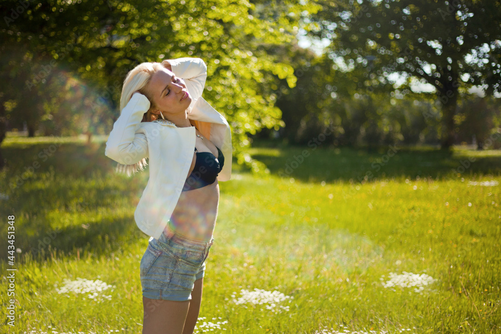 A teenage girl dressed in a jeans shorts and jacket straighting touching her hair in a city park. Woman portrait with a smile at sunny day at the summer.