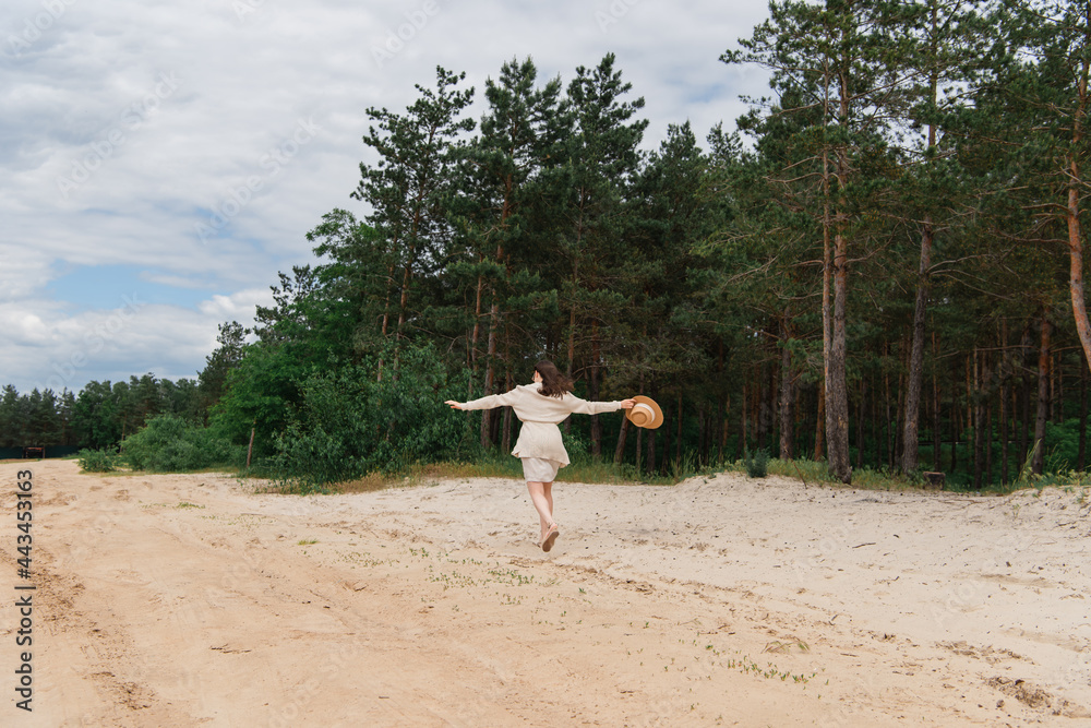 back view of young woman with outstretched hands holding straw hat and walking near forest back view of young woman with outstretched hands holding straw hat and walking near forest