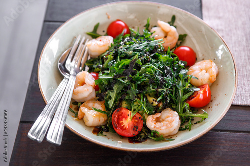 Salad of arugula, fresh tomatoes and shrimp, seasoned with sauce in a plate on a brown wooden board.