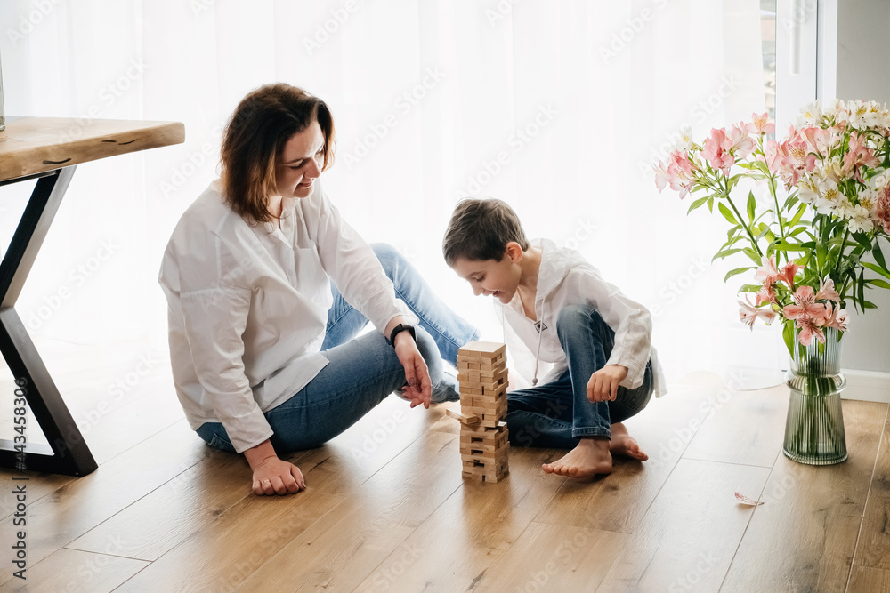 Mother with child son are sitting on the floor playing a board game of physical skill Tower. destroyed the tower. over sunny window. Jenga, janga of 54 wooden blocks. Popular family game at home