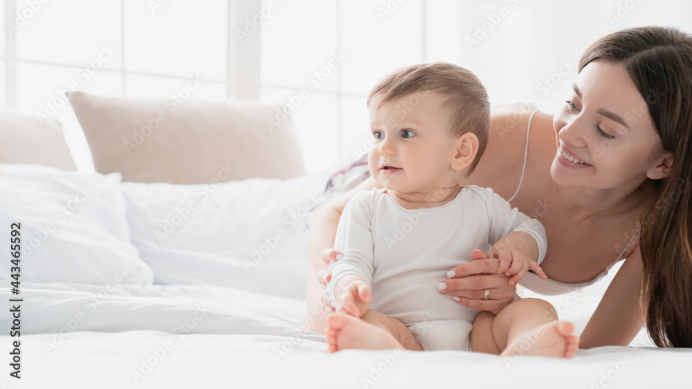 Lovely caucasian young mother playing with her little boy girl son daughter taking care of toddler infant newborn baby lying on the bed. Childcare and motherhood concept Lovely caucasian young mother playing with her little boy girl son daughter taking care of toddler infant newborn baby lying on the bed. Childcare and motherhood concept