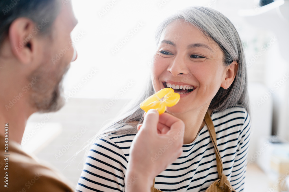 Happy mature husband man feeding his wife woman in the kitchen at home with bell pepper. Middle-aged family preparing cooking meal food together Happy mature husband man feeding his wife woman in the kitchen at home with bell pepper. Middle-aged family preparing cooking meal food together