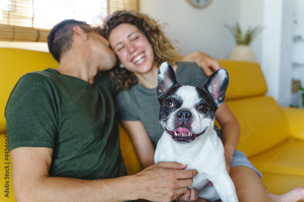Happy couple playing with dog at home. Horizontal view of couple laughing with bulldog pet on couch.