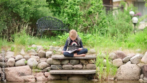 Cute little girl sitting cross-legged on stone steps near pond in backyard of house and reading book aloud. Child spending summer vacation in countryside photo