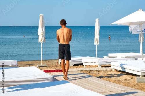 Young guy cleans the beach with a big brush mop. KOBLEVO, UKRAINE - JULY 7, 2020 photo