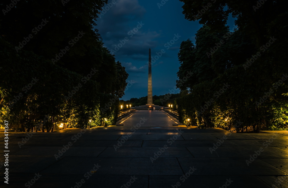 Monument to the Unknown Soldier with eternal fire in the evening. The tiled road in the night green park with lanterns. Illumination of a park road with lanterns. Park of Eternal Glory. Ukraine