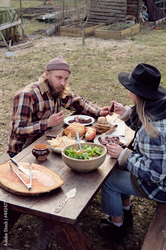 Young couple sitting at the table and eating fried meat with vegetables during barbecue outdoors