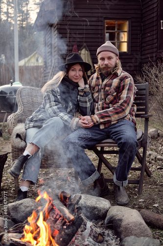 Portrait of young couple of farmers sitting on armchairs holding hands and looking at camera they resting near the fire outdoors